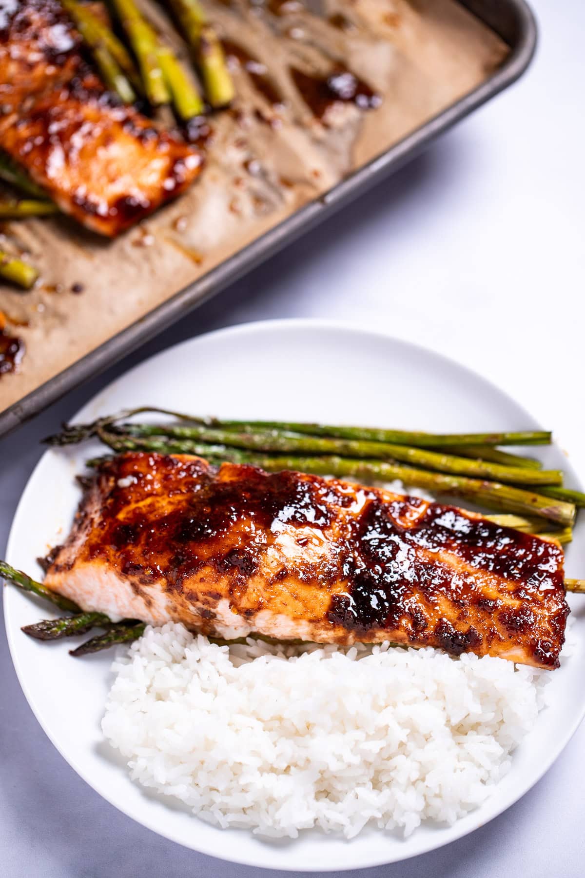 A plate with a balsamic glazed salmon fillet resting on top of rice and asparagus, with a sheet pan in the background.