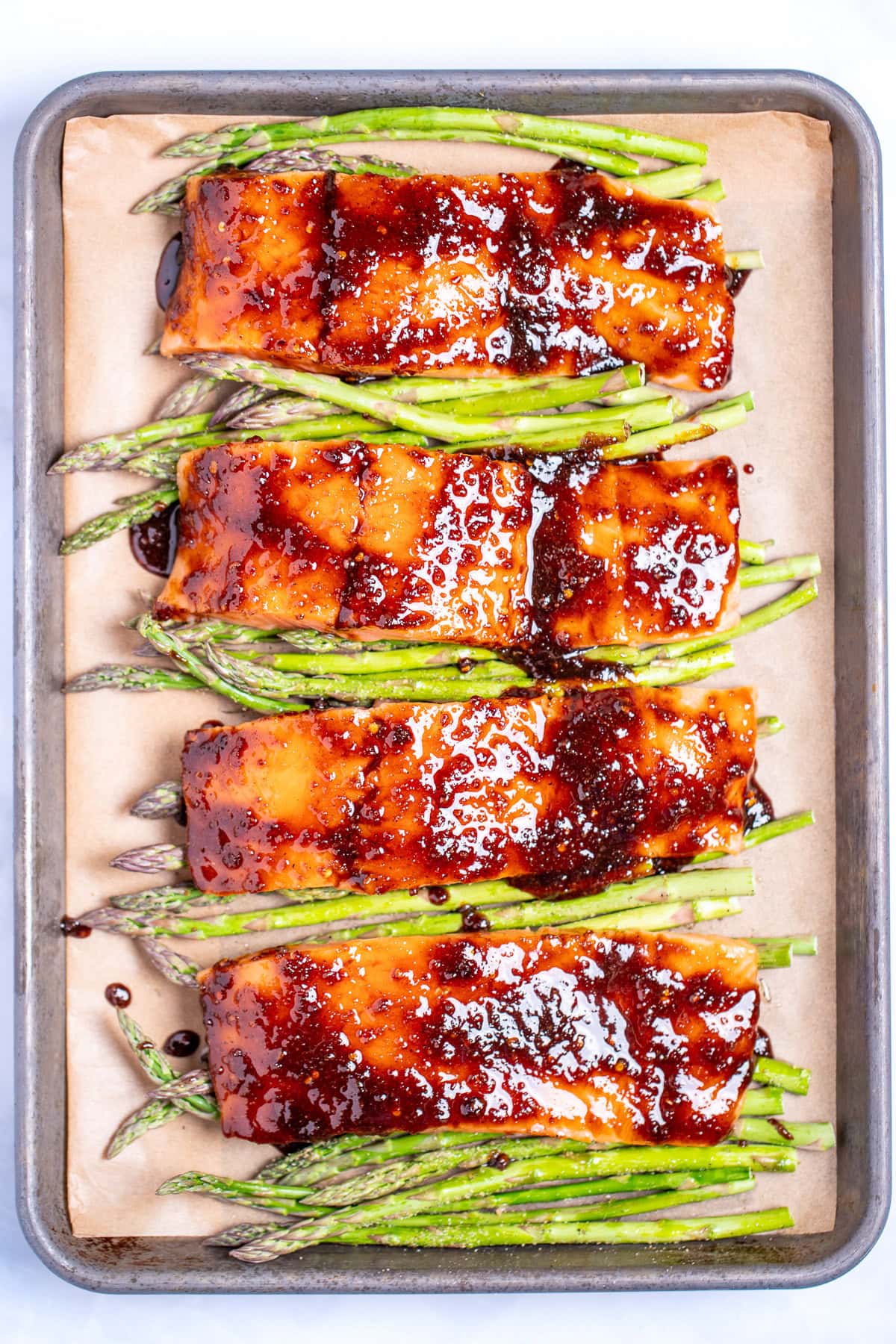 Four salmon fillets on a sheet pan lined with parchment paper, resting on top of asparagus, topped with a balsamic glaze, before being baked.