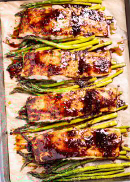 A sheet pan lined with parchment paper with four salmon fillets resting on top of asparagus, topped with a balsamic reduction.