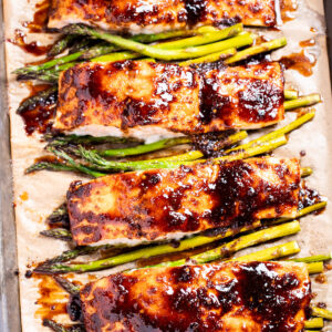 A sheet pan lined with parchment paper with four salmon fillets resting on top of asparagus, topped with a balsamic reduction.