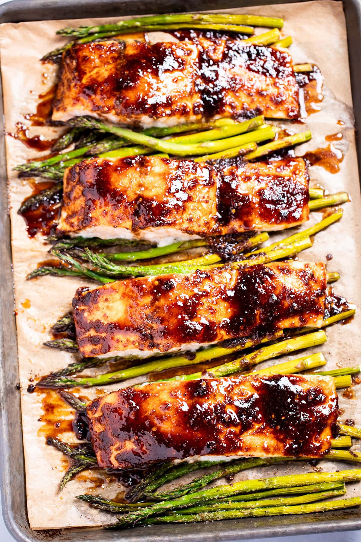 A sheet pan lined with parchment paper with four salmon fillets resting on top of asparagus, topped with a balsamic reduction.