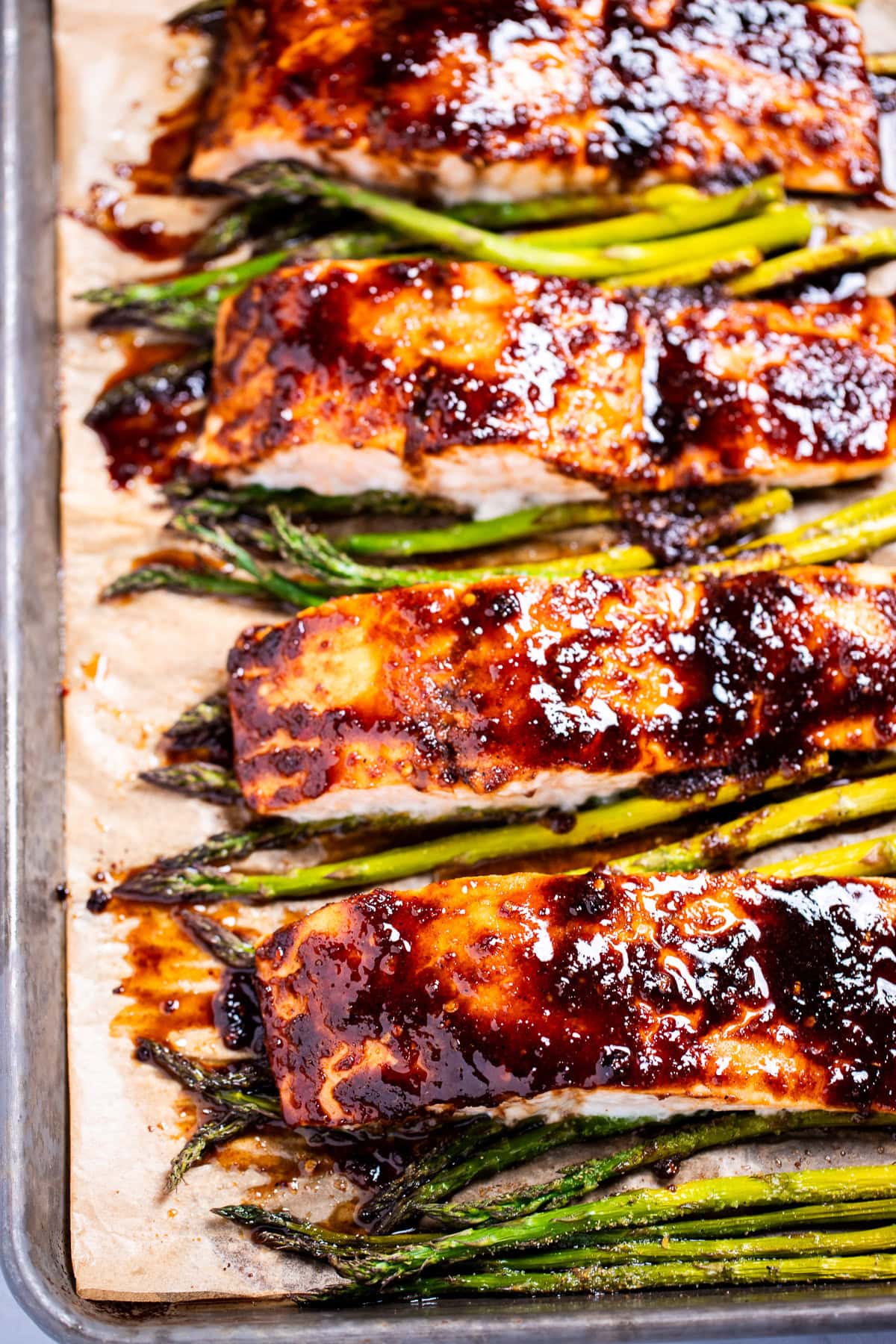 A sheet pan lined with parchment paper with four salmon fillets resting on top of asparagus, topped with a balsamic reduction.