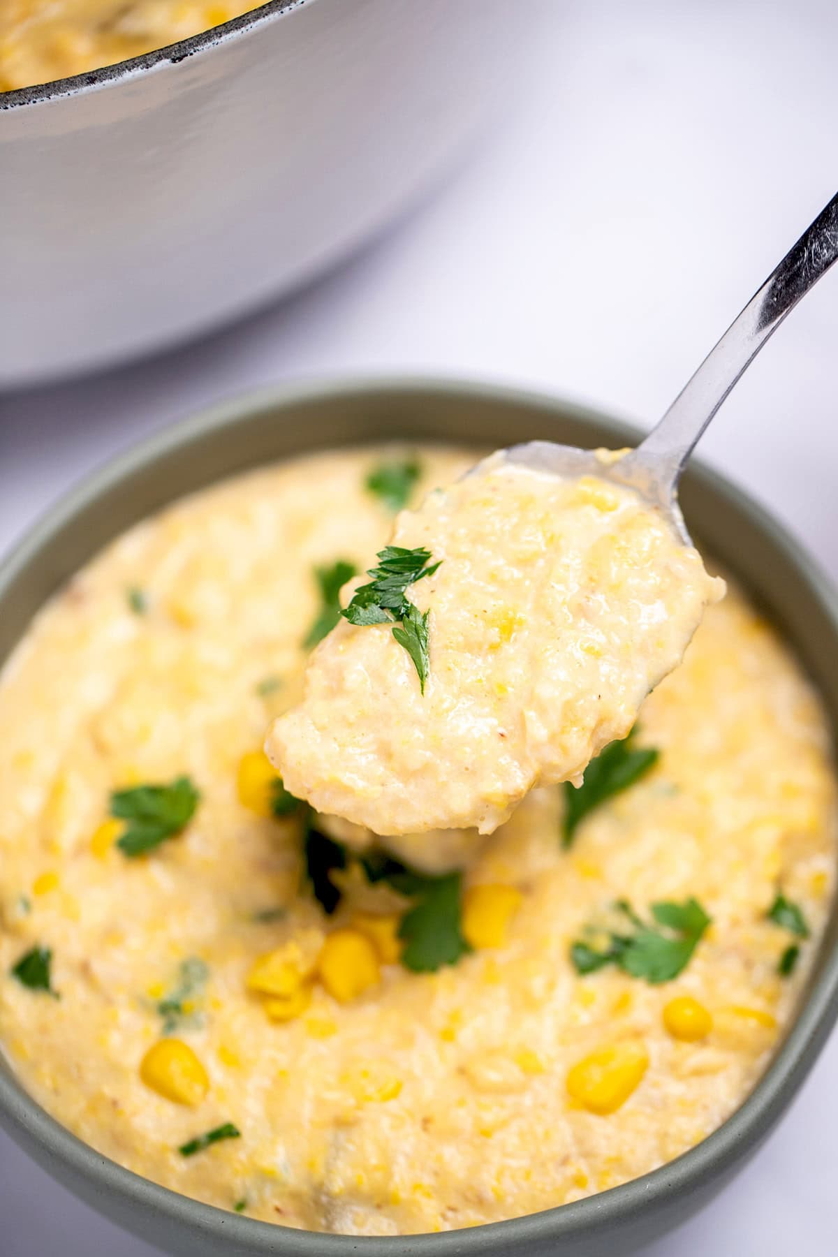 A bowl sitting on the table with crab and corn chowder topped with corn kernels and fresh parsley, with a spoon lifting some up, and a dutch oven in the background.