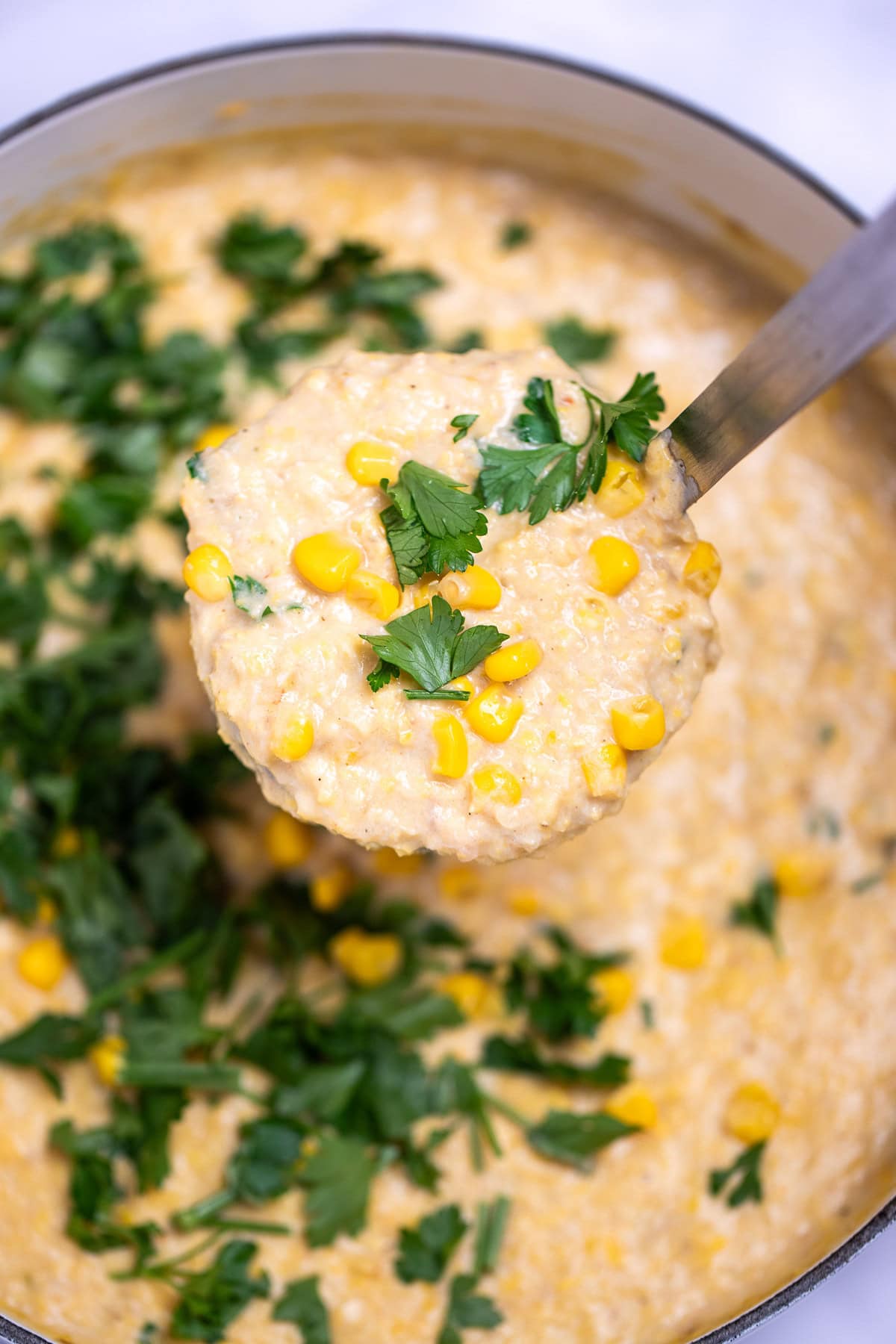 A dutch oven of crab and corn chowder topped with corn kernels and fresh parsley, with a ladle lifting some chowder up.