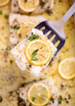A baking dish with four cooked cod fillets, topped with lemon slices and fresh parsley, and in a lemon butter sauce. A spatula is lifting up one of the cod pieces.
