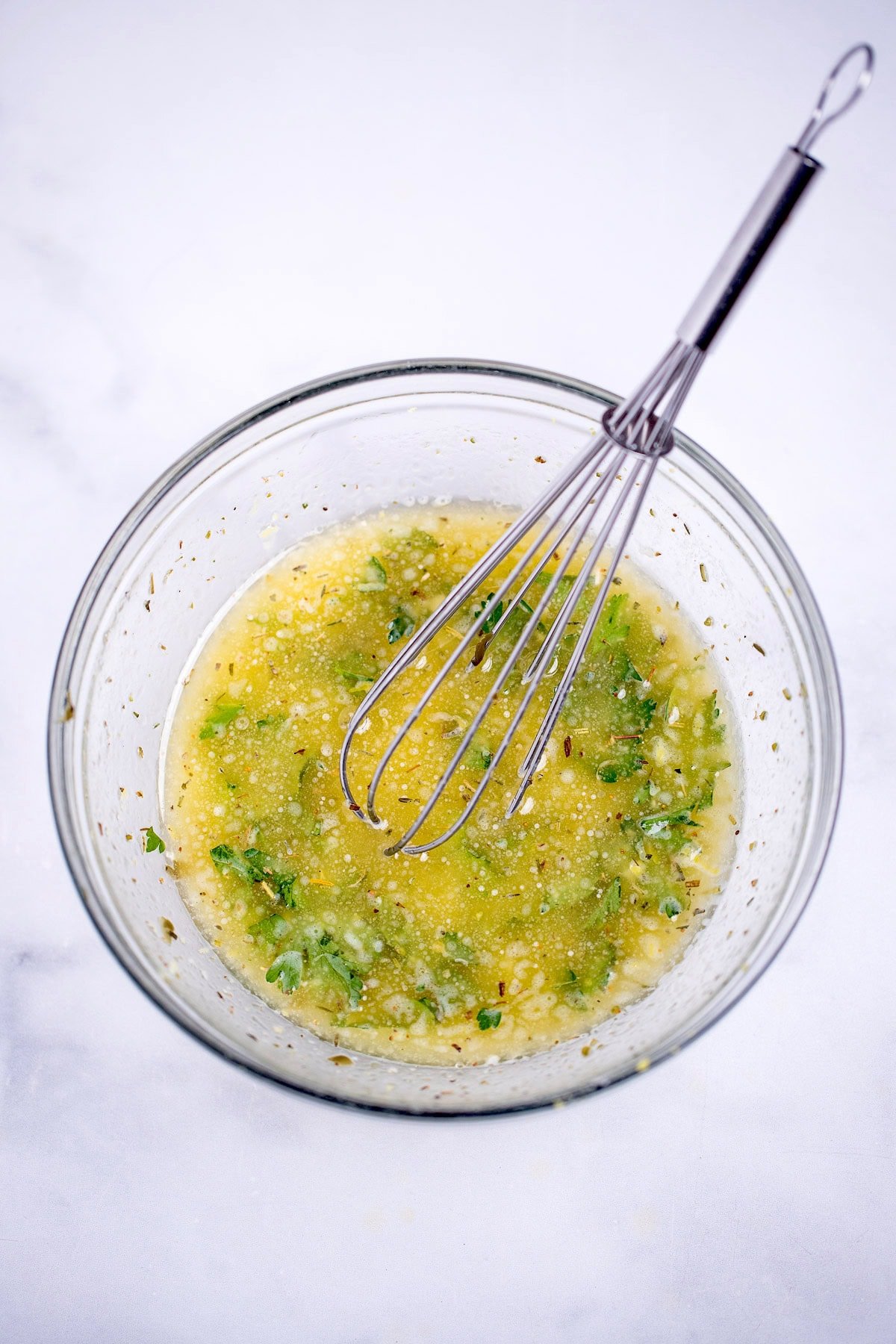 A small glass bowl with melted butter whisked with capers, parsley, garlic, and seasoning with a whisk in the bowl, resting on a table.