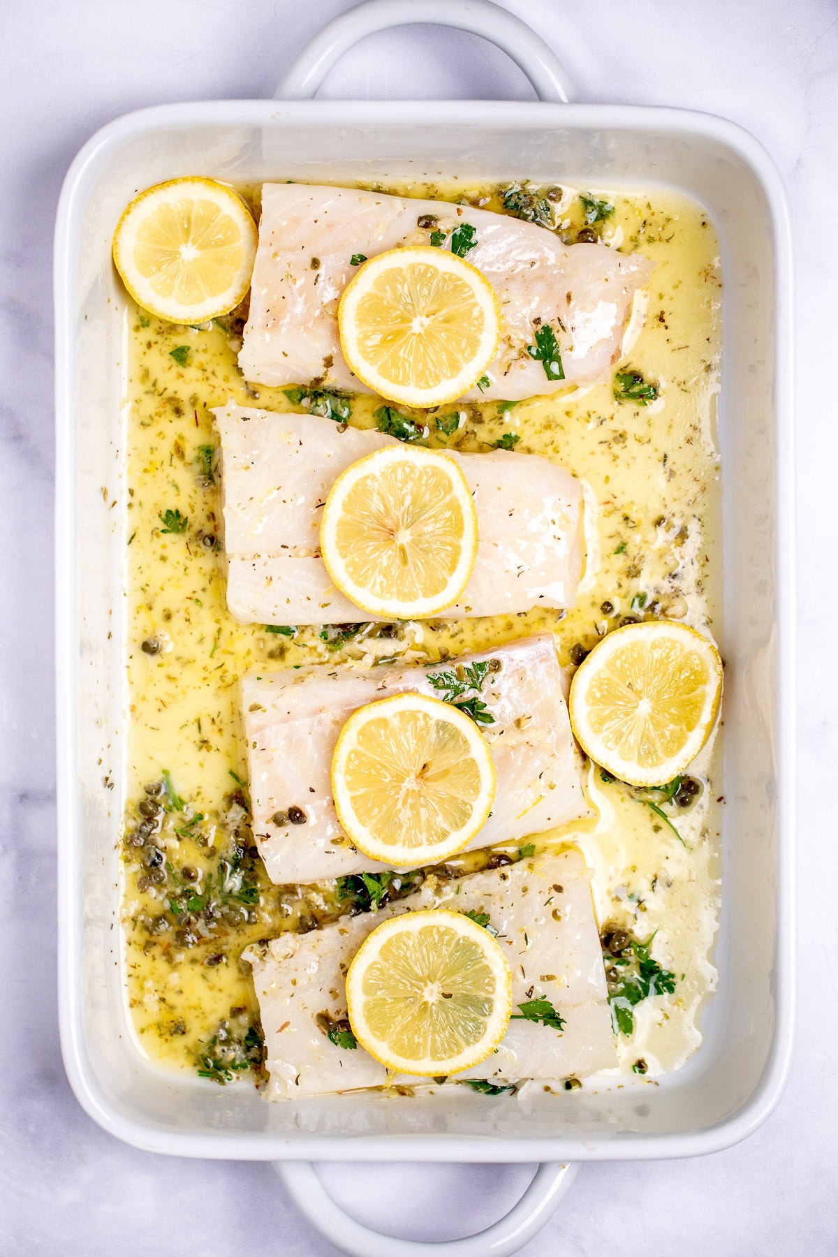 A baking dish with raw cod fillets in a lemon butter sauce topped with lemon slices before being cooked.