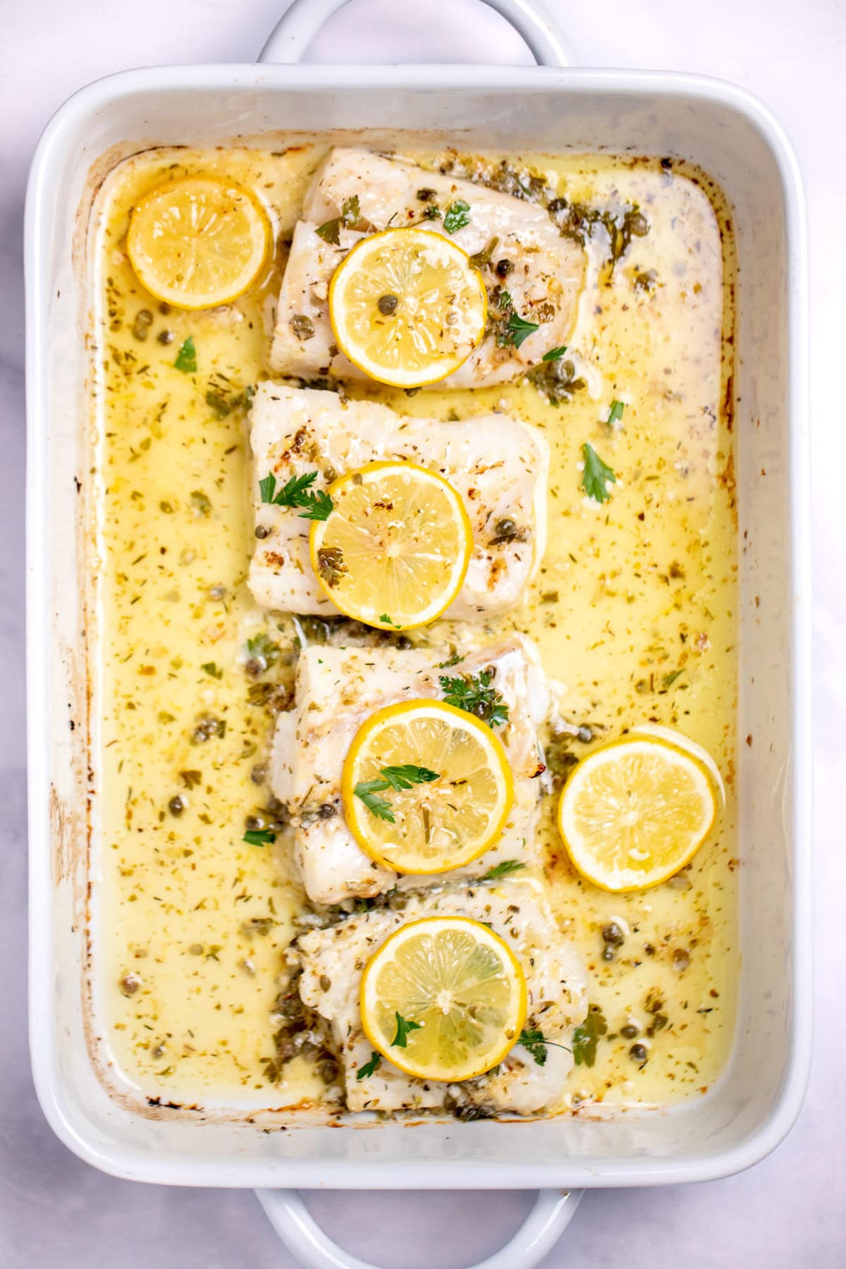 A baking dish on a table, with four cooked cod fillets, topped with lemon slices and fresh parsley, and in a lemon butter sauce.