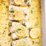 A baking dish on a table, with four cooked cod fillets, topped with lemon slices and fresh parsley, and in a lemon butter sauce.