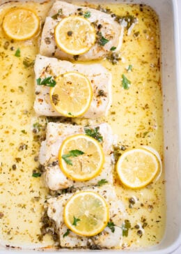 A baking dish on a table, with four cooked cod fillets, topped with lemon slices and fresh parsley, and in a lemon butter sauce.