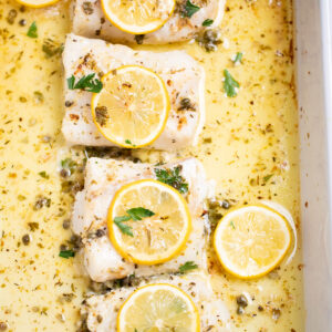 A baking dish on a table, with four cooked cod fillets, topped with lemon slices and fresh parsley, and in a lemon butter sauce.