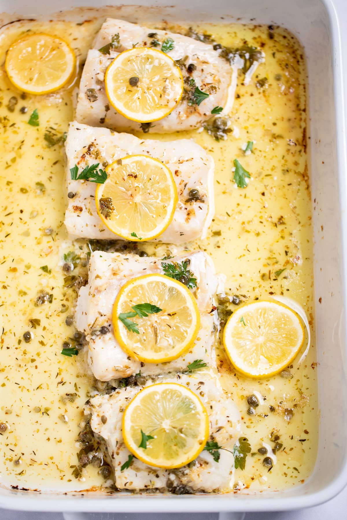 A baking dish on a table, with four cooked cod fillets, topped with lemon slices and fresh parsley, and in a lemon butter sauce.