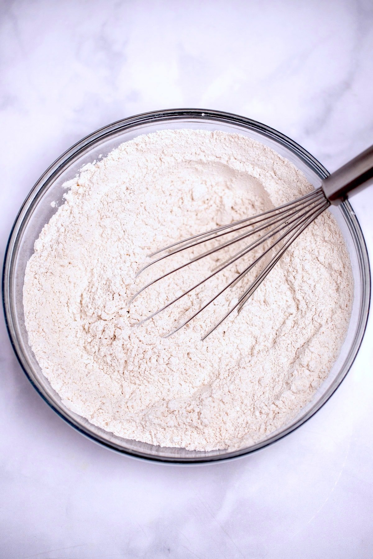 A glass bowl of flour with a whisk on a table.