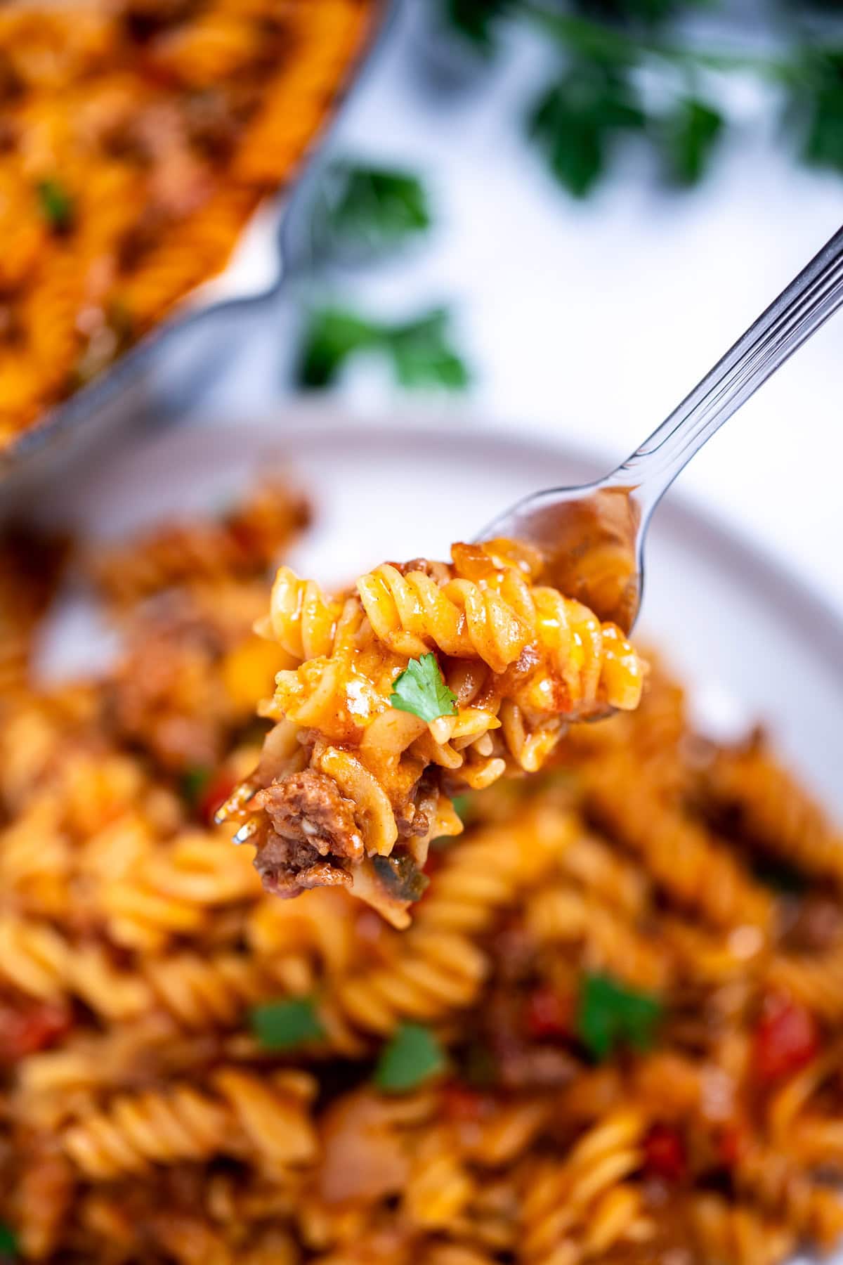 A fork holding pasta and ground beef topped with fresh parsley, over a plate of cheeseburger casserole, with the casserole skillet and fresh parsley in the background.