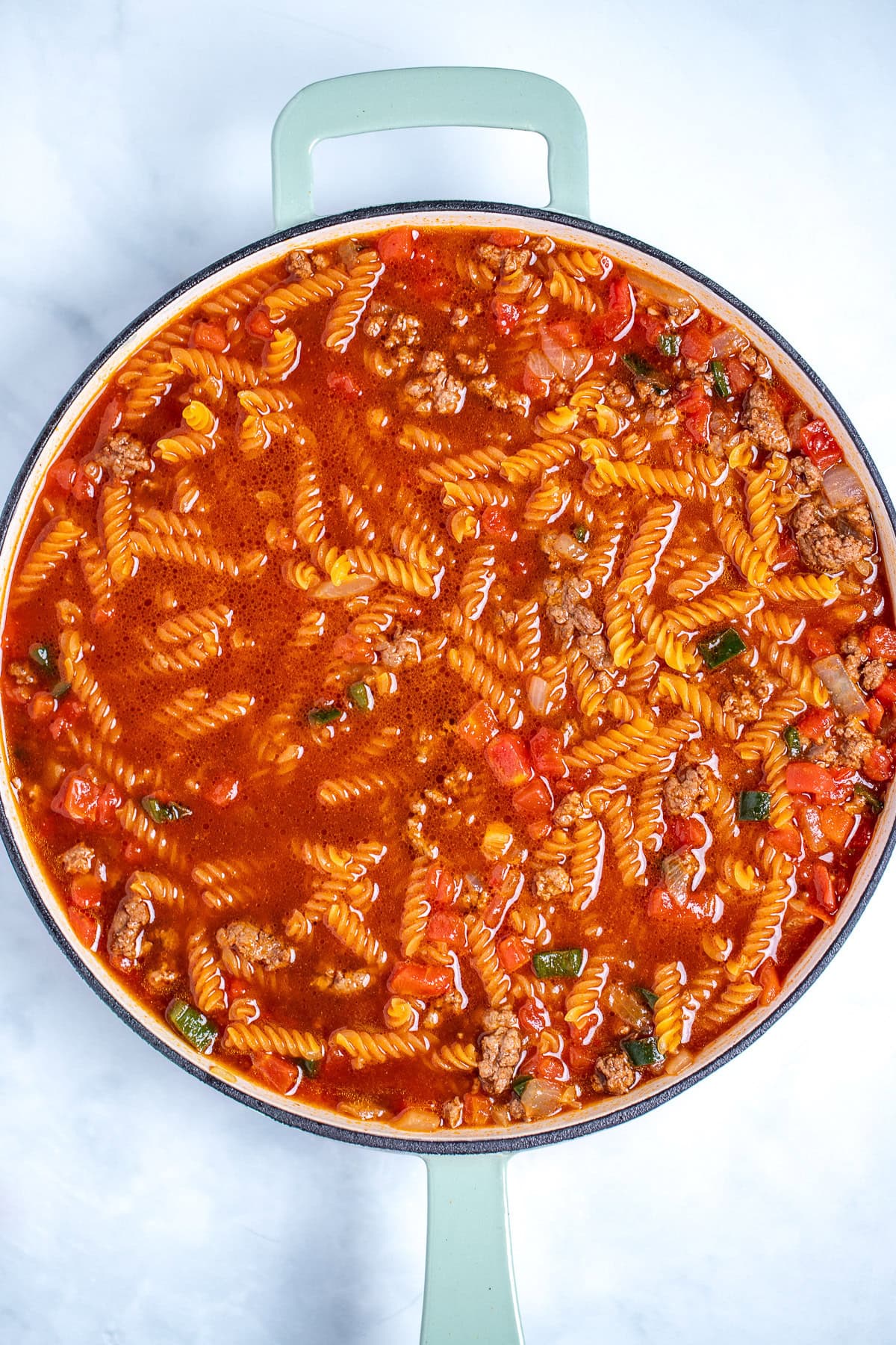 A skillet with ground beef, broth, and tomato sauce with pasta before cooking.