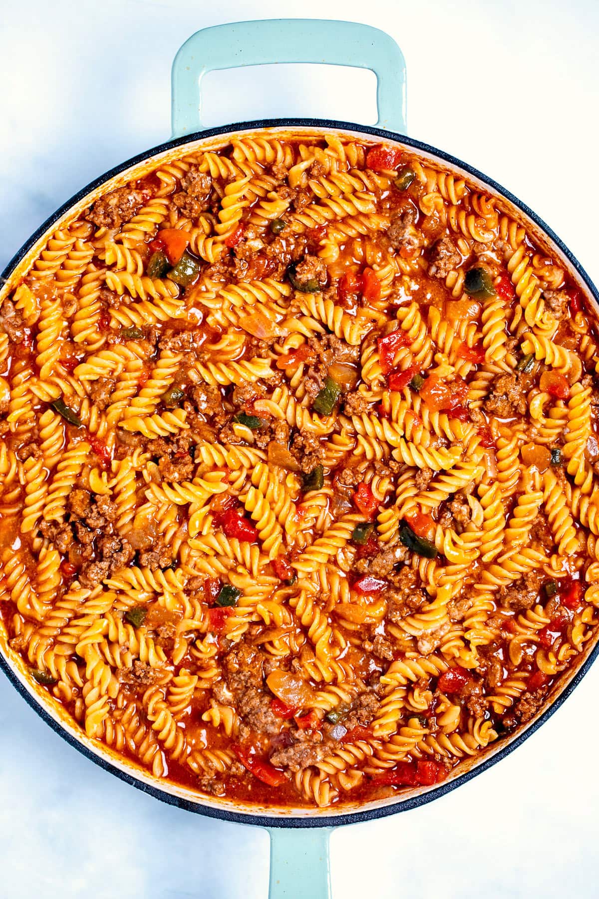 A skillet with pasta, onion, peppers, and ground beef cooked in broth and tomato sauce, before topping it with cheese.
