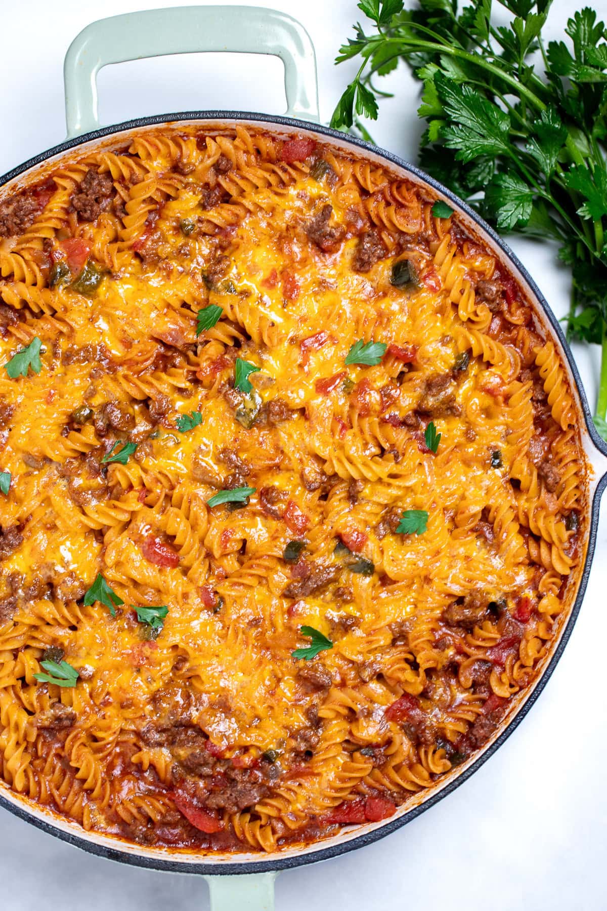 A cheeseburger casserole skillet with ground beef, gluten free rotini pasta, peppers, and onion topped with cheddar cheese and broiled, then topped with fresh parsley. There's fresh parsley on the table next to the skillet.