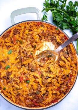 A cheeseburger casserole skillet with ground beef, gluten free rotini pasta, peppers, and onion topped with cheddar cheese and broiled, then topped with fresh parsley. There's a spoon in the skillet after having scooped some out, and fresh parsley on the table next to the skillet.