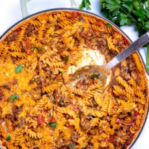 A cheeseburger casserole skillet with ground beef, gluten free rotini pasta, peppers, and onion topped with cheddar cheese and broiled, then topped with fresh parsley. There's a spoon in the skillet after having scooped some out, and fresh parsley on the table next to the skillet.