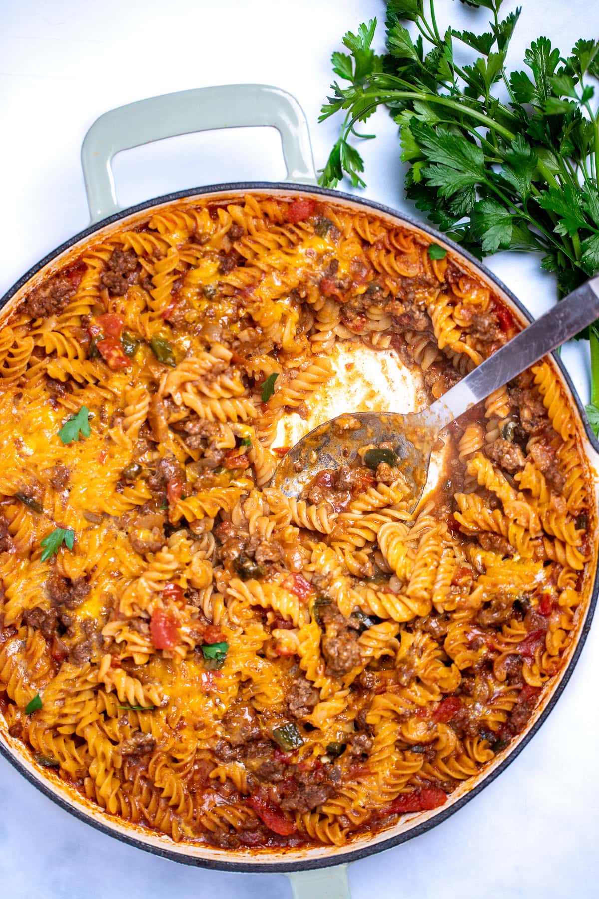 A cheeseburger casserole skillet with ground beef, gluten free rotini pasta, peppers, and onion topped with cheddar cheese and broiled, then topped with fresh parsley. There's a spoon in the skillet after having scooped some out, and fresh parsley on the table next to the skillet.
