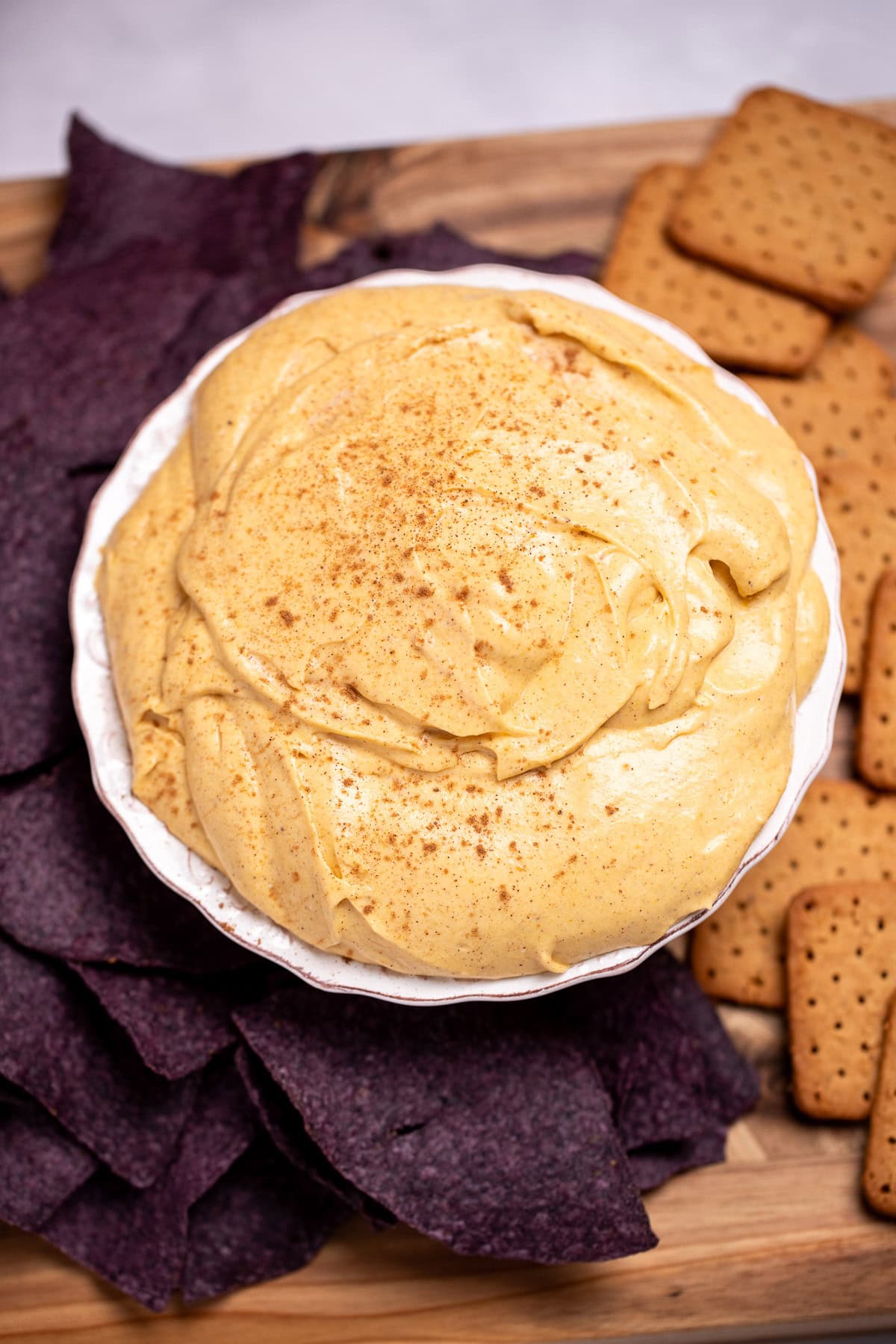 A bowl full of pumpkin pie dip resting on a cutting board, surrounded by gluten free graham crackers and blue corn chips.