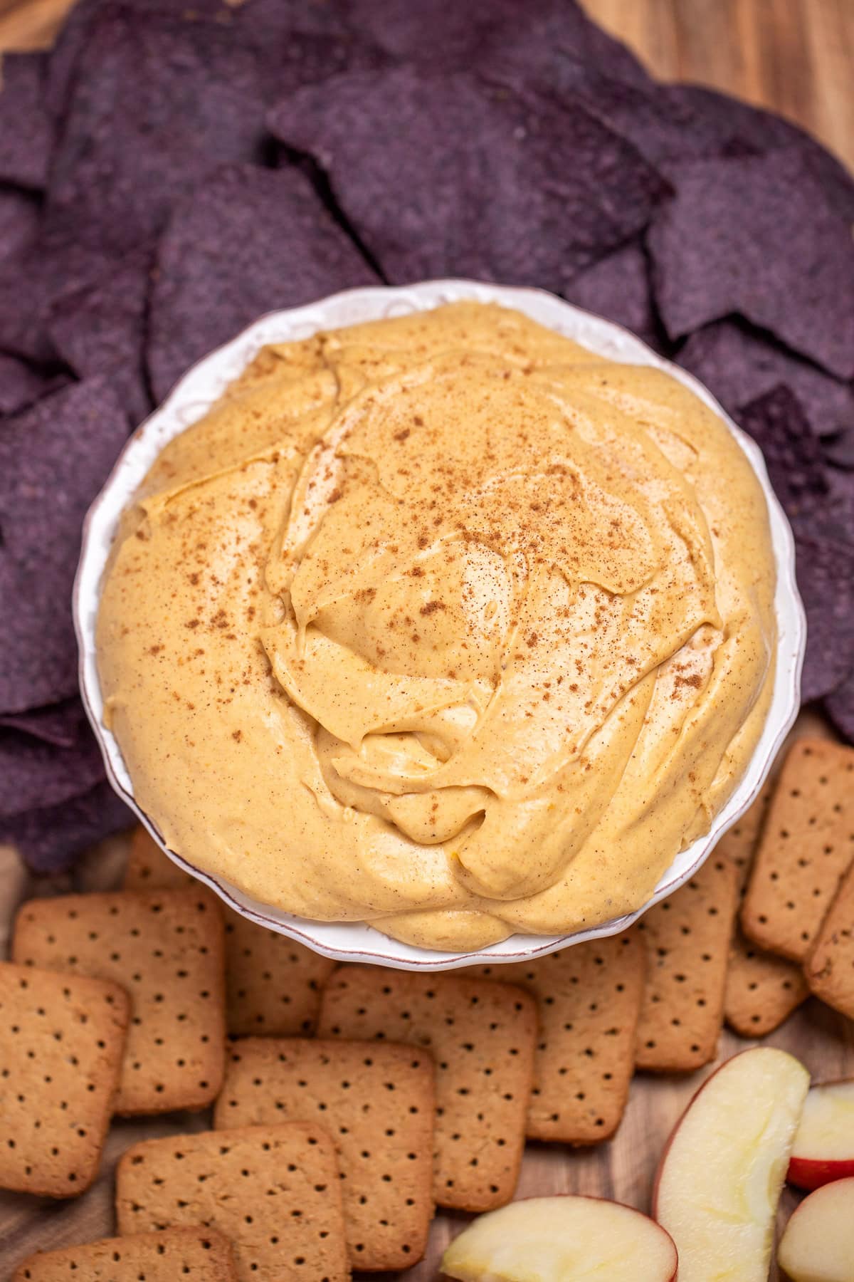 A bowl full of pumpkin pie dip resting on a cutting board, surrounded by gluten free graham crackers, apple slices, and blue corn chips.