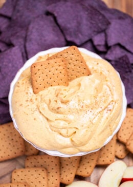 A bowl full of pumpkin pie dip resting on a cutting board, surrounded by gluten free graham crackers, apple slices, and blue corn chips. Two graham crackers are resting in the dip.