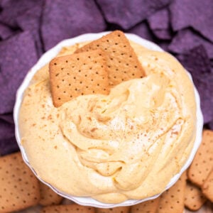 A bowl full of pumpkin pie dip resting on a cutting board, surrounded by gluten free graham crackers, apple slices, and blue corn chips. Two graham crackers are resting in the dip.