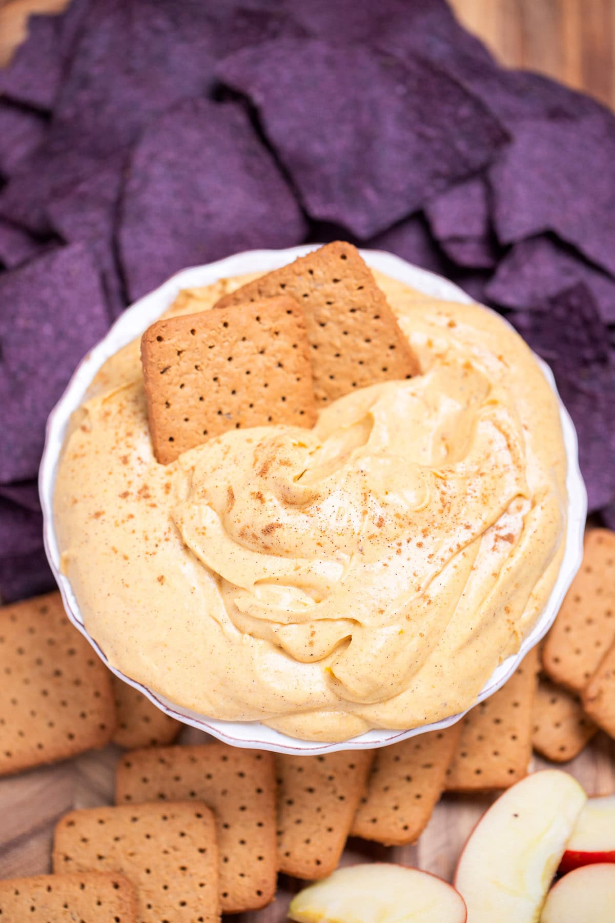 A bowl full of pumpkin pie dip resting on a cutting board, surrounded by gluten free graham crackers, apple slices, and blue corn chips. Two graham crackers are resting in the dip.