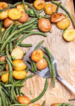 Pinterest pin with a sheet pan lined with parchment paper with roasted green beans and potatoes with olive oil and spices, topped with fresh parsley, and a spatula resting on the sheet pan.