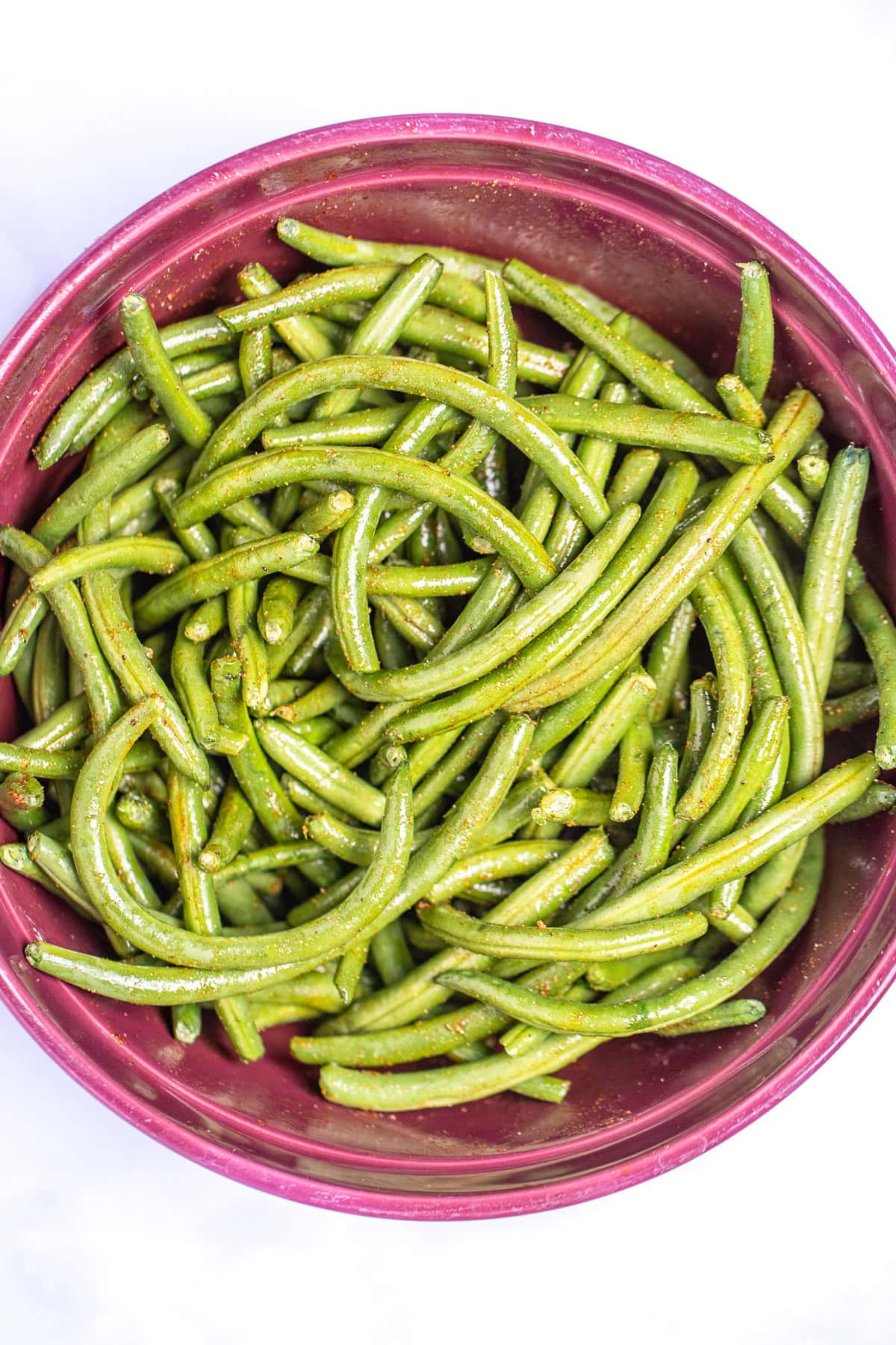 A mixing bowl with green beans tossed with olive oil and seasoning before being cooked.