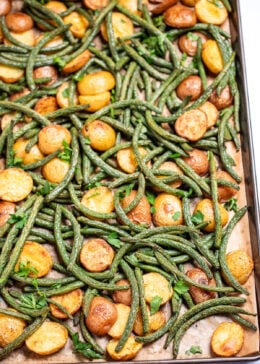 A sheet pan lined with parchment paper with roasted green beans and potatoes with olive oil and spices, topped with fresh parsley.