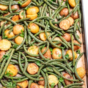 A sheet pan lined with parchment paper with roasted green beans and potatoes with olive oil and spices, topped with fresh parsley.