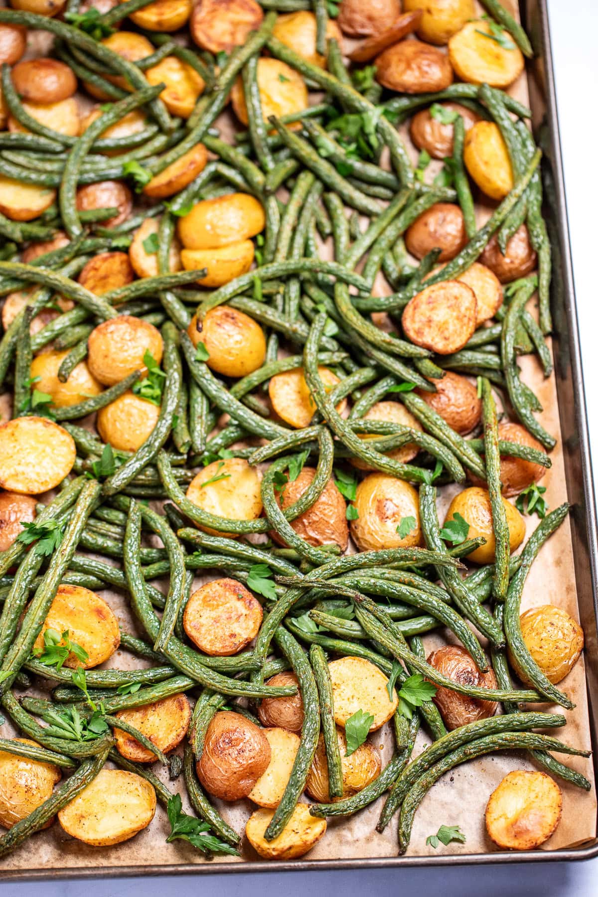 A sheet pan lined with parchment paper with roasted green beans and potatoes with olive oil and spices, topped with fresh parsley.