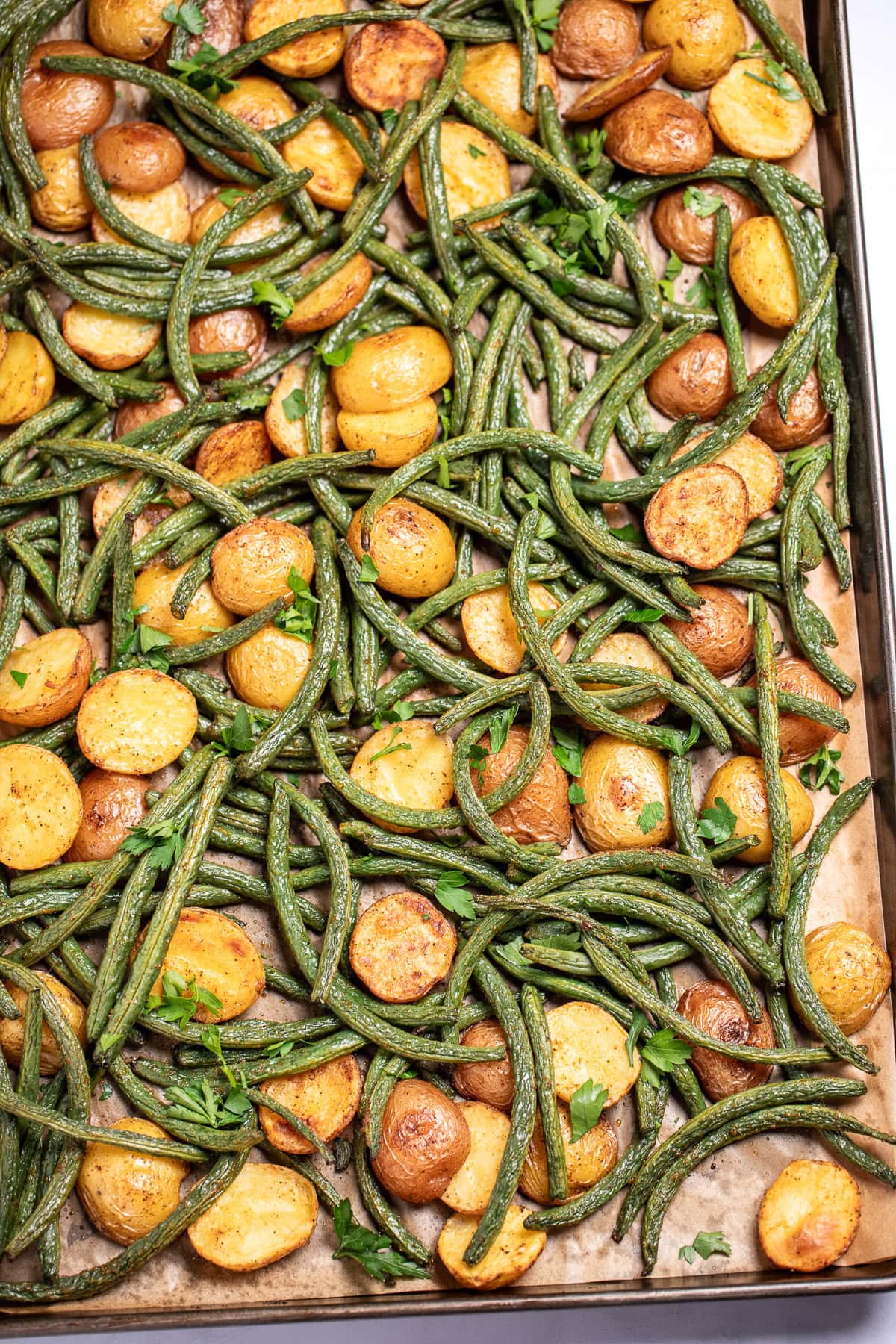 A sheet pan lined with parchment paper with roasted green beans and potatoes with olive oil and spices, topped with fresh parsley.
