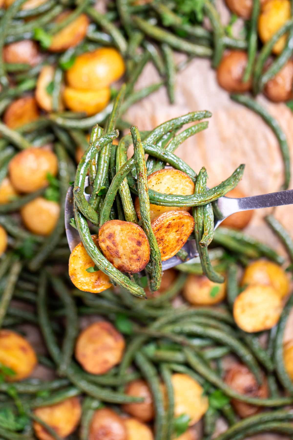 A sheet pan lined with parchment paper with roasted green beans and potatoes with olive oil and spices, topped with fresh parsley, and a spatula lifting up some potatoes and green beans.