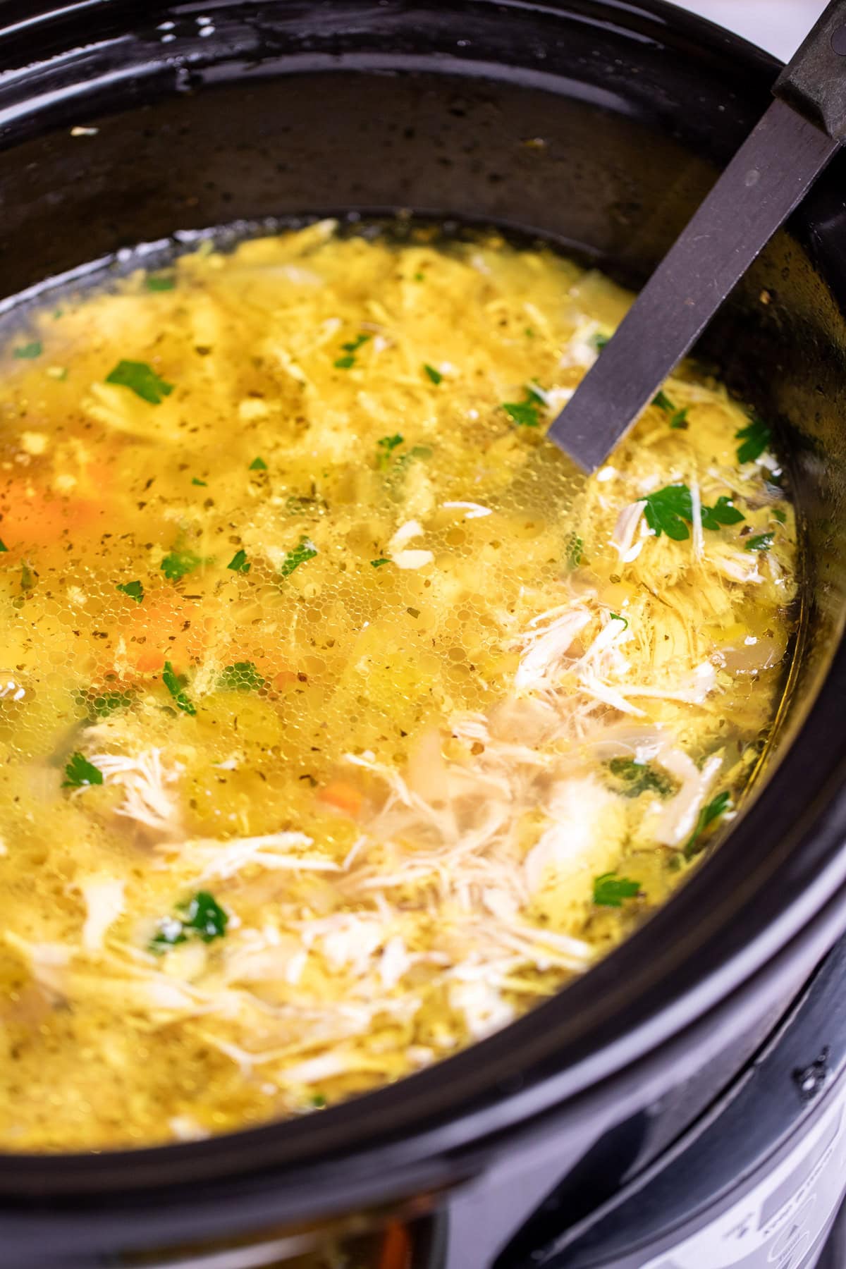 A slow cooker of chicken soup topped with fresh parsley with a ladle in it ready to serve.