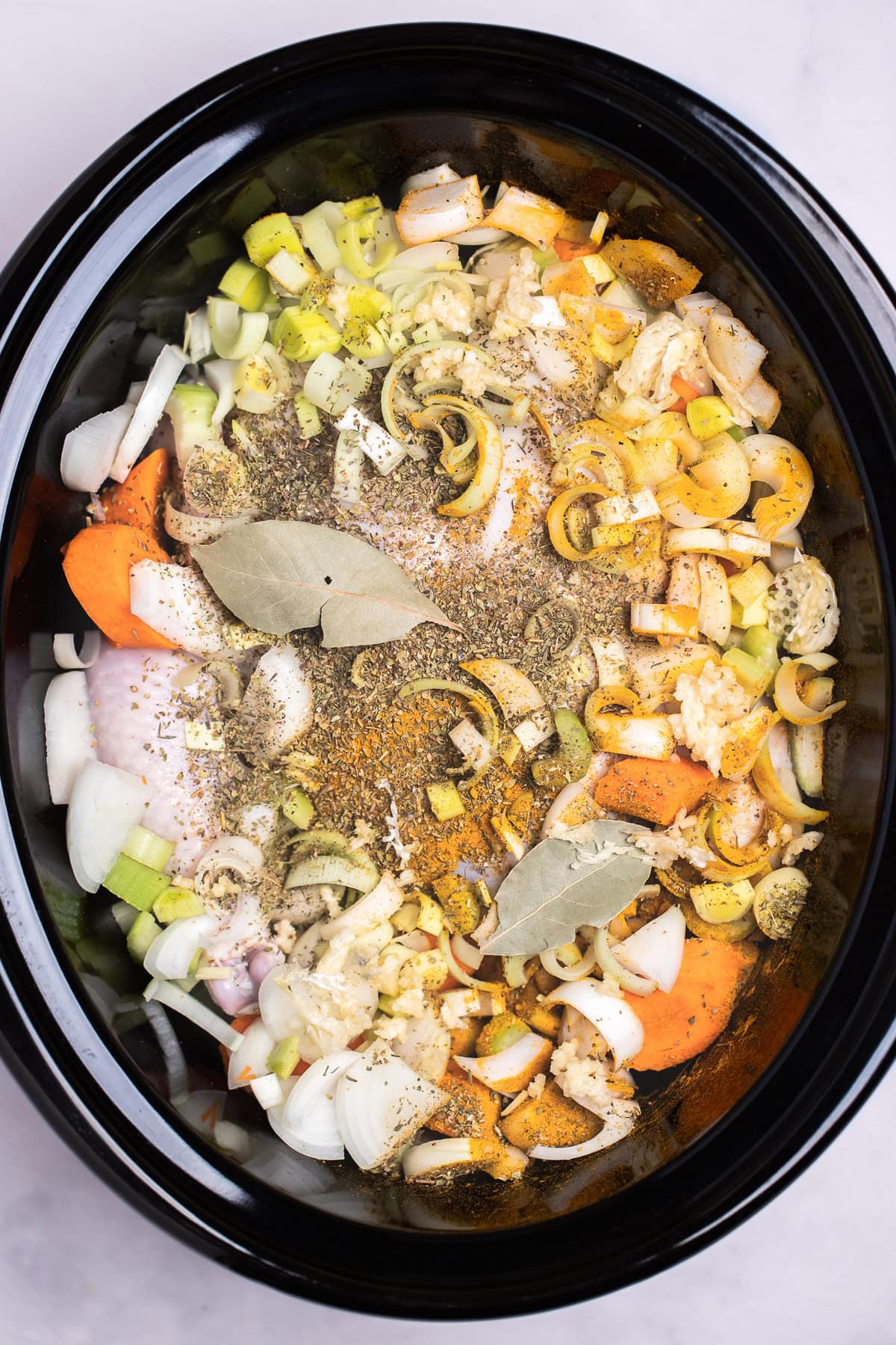 A slow cooker pot with a raw whole chicken topped with vegetables and spices, before adding the water and cooking.