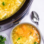 A bowl of chicken soup on a table, next to a smaller bowl of fresh parsley to the left and a spoon on the table to the right, in front of a slow cooker of soup.