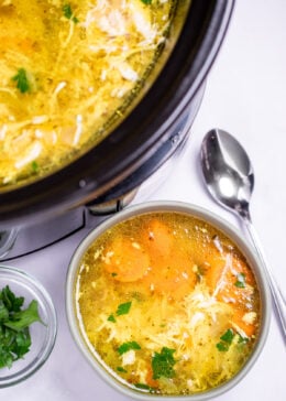 A bowl of chicken soup on a table, next to a smaller bowl of fresh parsley to the left and a spoon on the table to the right, in front of a slow cooker of soup.