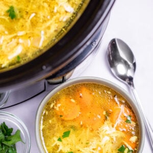 A bowl of chicken soup on a table, next to a smaller bowl of fresh parsley to the left and a spoon on the table to the right, in front of a slow cooker of soup.