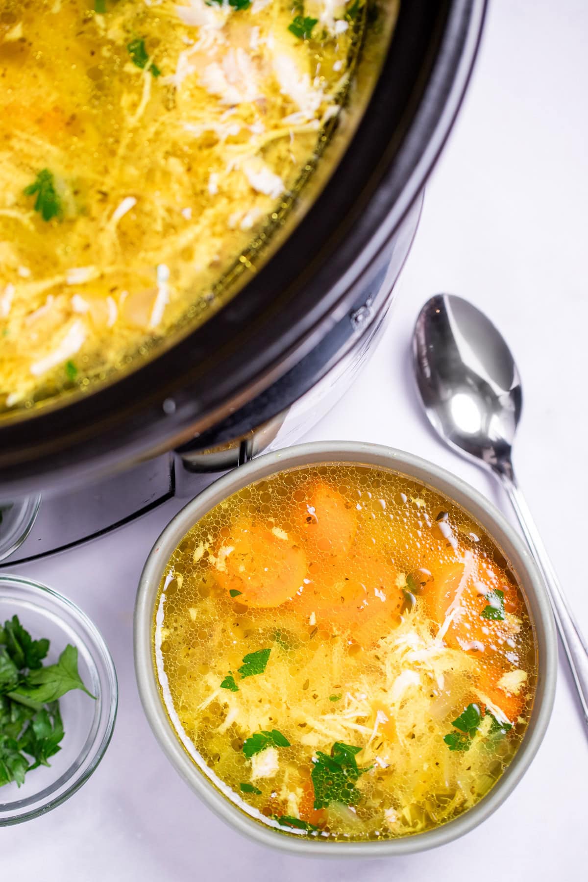 A bowl of chicken soup on a table, next to a smaller bowl of fresh parsley to the left and a spoon on the table to the right, in front of a slow cooker of soup.