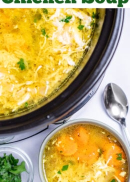 Pinterest pin with a bowl of chicken soup on a table, next to a smaller bowl of fresh parsley to the left and a spoon on the table to the right, in front of a slow cooker of soup.