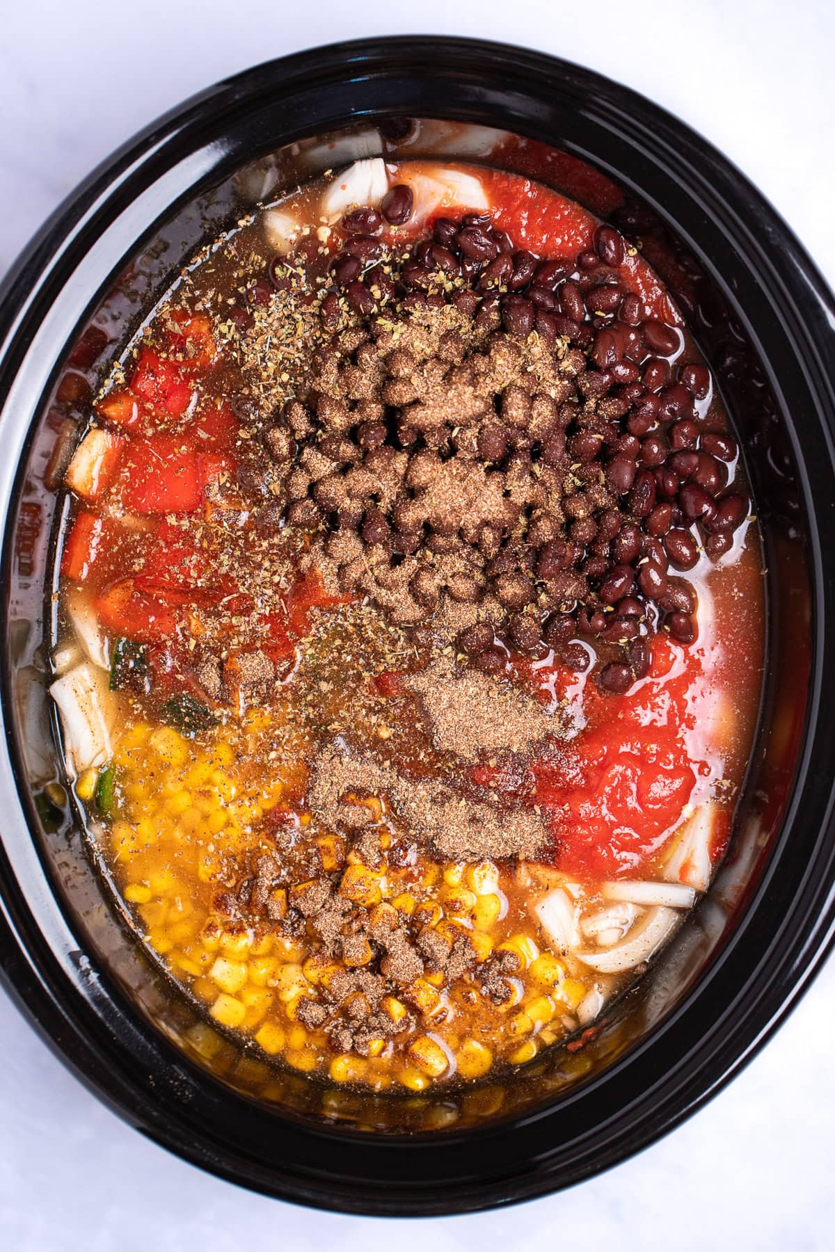 A slow cooker pot with corn, black beans, crushed tomatoes, chopped onion, broth, and seasoning before being cooked.