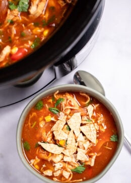 A bowl of chicken tortilla soup topped with fresh cilantro and crushed tortilla chips with a spoon on the table next to it, and the crockpot behind it.