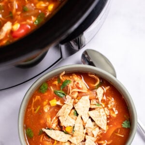 A bowl of chicken tortilla soup topped with fresh cilantro and crushed tortilla chips with a spoon on the table next to it, and the crockpot behind it.