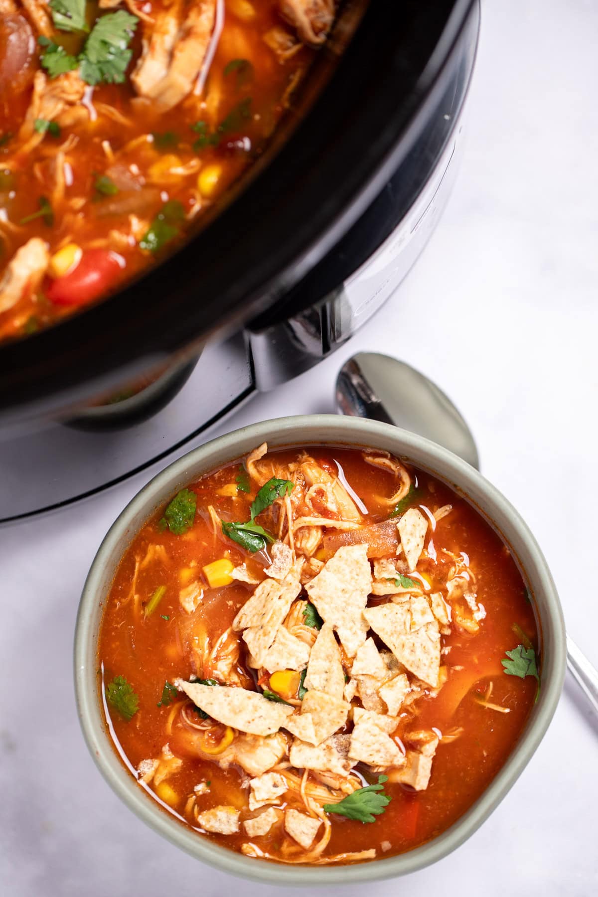 A bowl of chicken tortilla soup topped with fresh cilantro and crushed tortilla chips with a spoon on the table next to it, and the crockpot behind it.