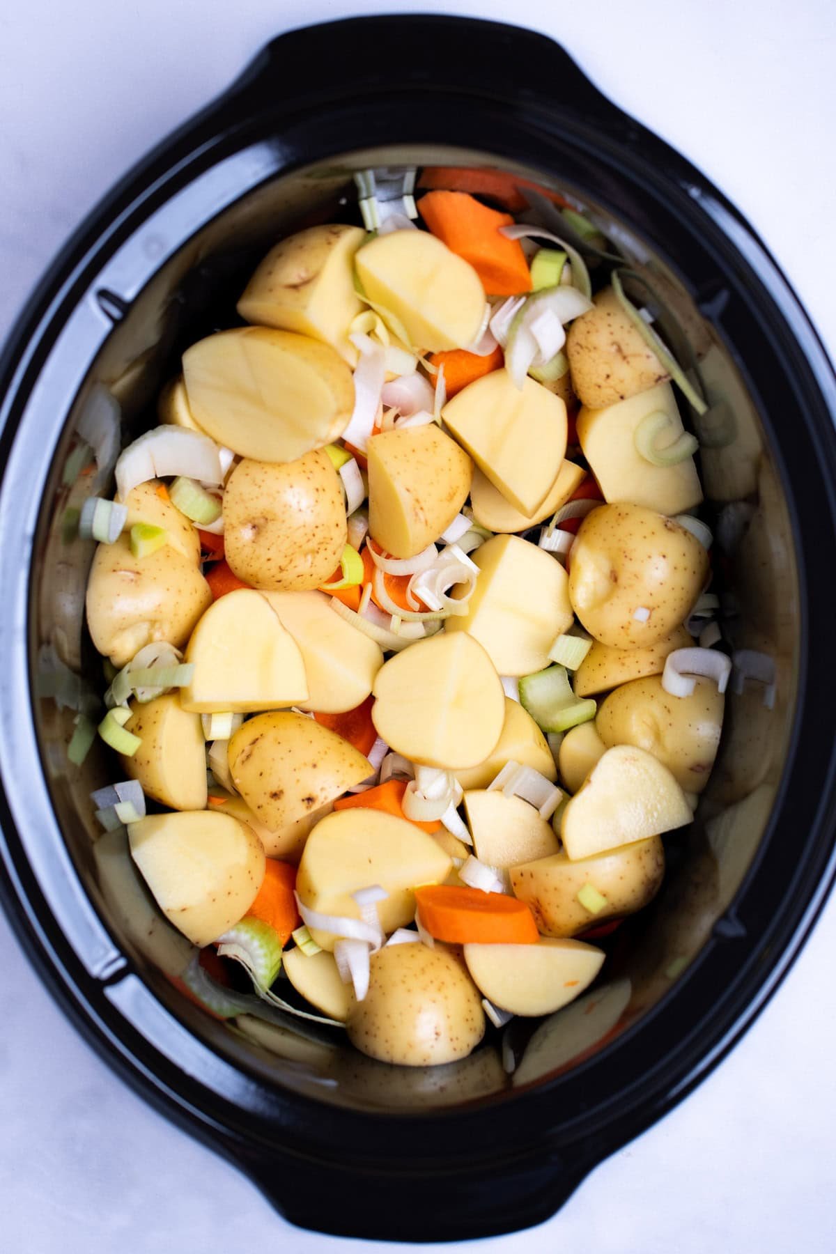 A slow cooker with quartered potatoes, carrots, and leeks before being cooked.