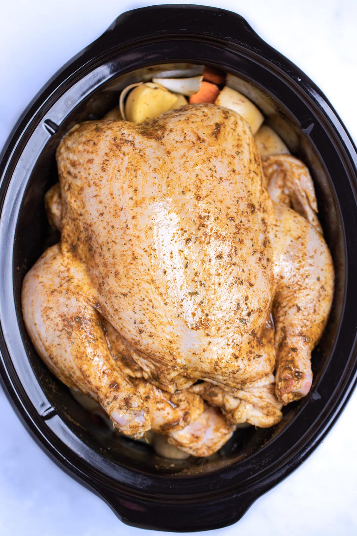 A slow cooker with a raw chicken rubbed with spices and olive oil, resting on top of potatoes, before being cooked.