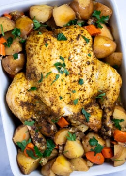 A whole chicken, broiled until crispy, resting on top of potatoes and carrots in a baking dish, topped with fresh parsley.